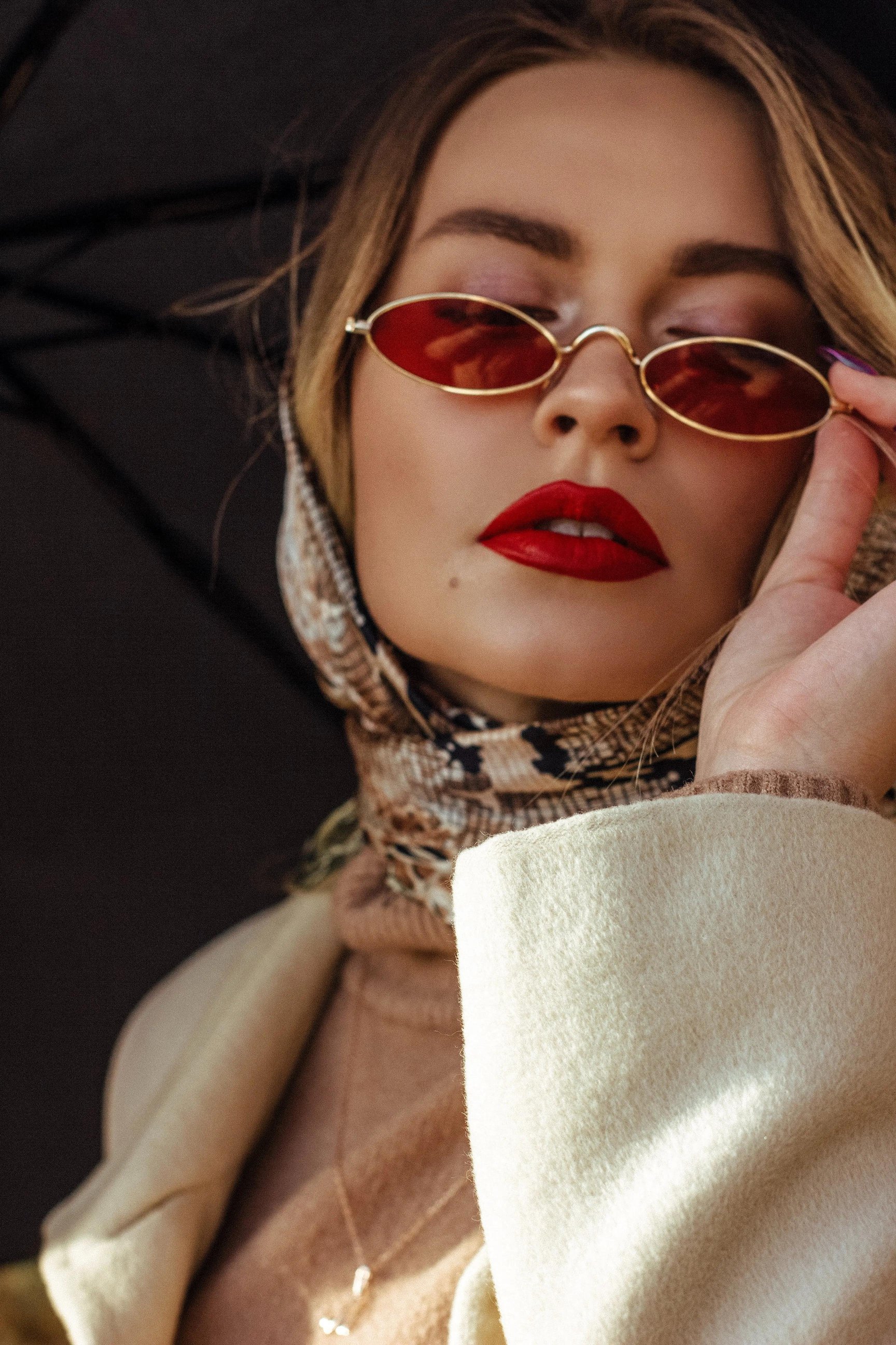 a woman wearing red lipstick and sunglasses with a scarf holding her sunglass